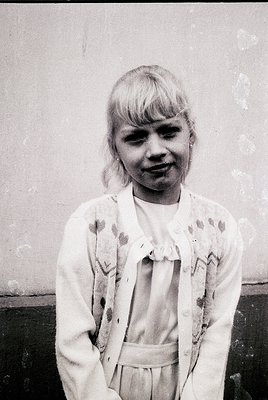 Young girl in 1960s-70s fashion, posing against a plain wall. She wears a floral-patterned blouse with a belted cardigan, sho...