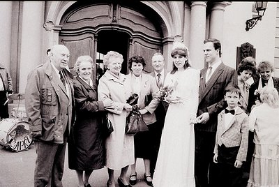Black-and-white wedding photo outside a grand, arched entrance with classical columns. A bride in a simple white gown holds f...