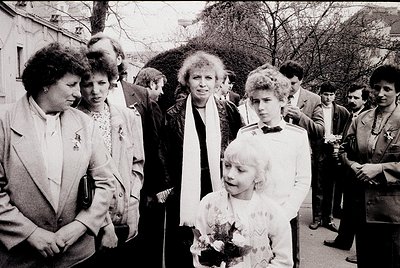 Black-and-white street scene featuring a group of adults and children in formal attire, likely 1970s–1980s. Central figure in...