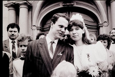 Classic black-and-white wedding photo featuring a bride in a simple white gown with veil and bouquet, posing with groom in a ...