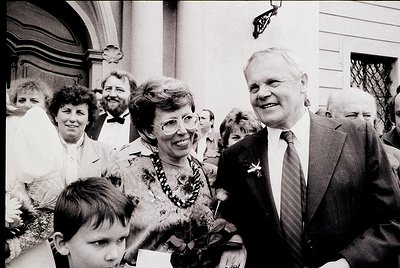 Black-and-white formal gathering featuring a man in a suit with a floral boutonnière greeting a woman in a floral dress, surr...