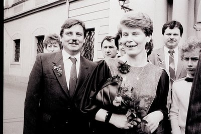 Couple in formal attire at what appears to be a wedding, holding floral bouquets. Man in suit with boutonnière, woman in knee...