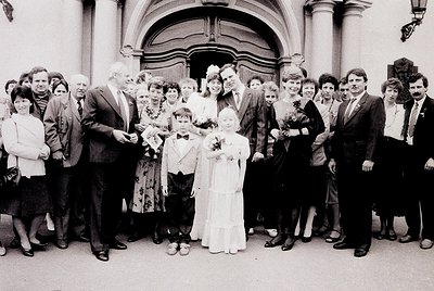 A black-and-white wedding photo featuring a bride in a classic A-line gown and groom in a suit, flanked by 16 guests in 1970s...