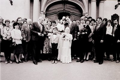 Family wedding photo outside a grand, arched entrance, likely 1970s–1980s. Bride in white gown with bouquet, groom in suit, s...