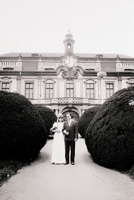 Classic 1960s-70s wedding portrait in grand European courtyard. Couple in formal attire—bride in floor-length gown, groom in ...