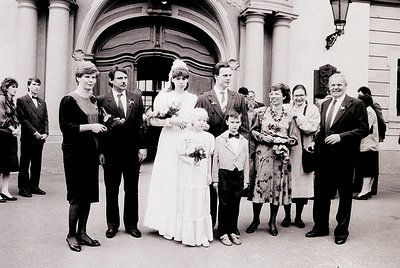 Neatly posed wedding group photo in front of a grand, arched entrance with classical columns. Bride in simple white gown with...