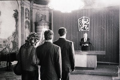 Black-and-white formal event in ornate hall featuring three men in suits and a woman in a veil, facing a podium with a coat-o...