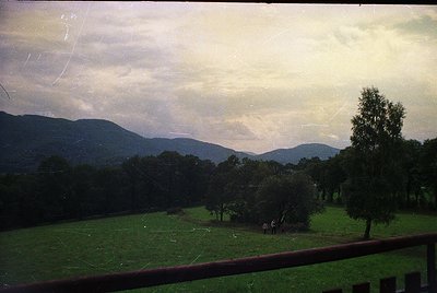 Vintage landscape shot through a window frame, showcasing rolling hills and dense forest under overcast skies. Two figures st...
