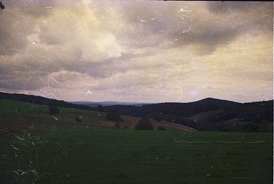 Vintage landscape photograph featuring rolling hills under dramatic, textured clouds. Evidence of aging film grain and slight...