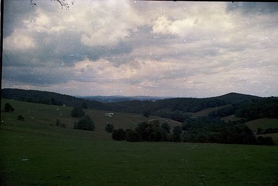 Vintage landscape shot of rolling hills under dramatic cloud cover, likely mid-20th century. Green pastures with scattered tr...