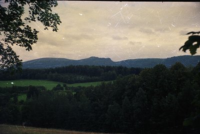 Vintage slide of lush valley framed by dense forest and rolling hills under soft, diffused light. Green meadows contrast with...