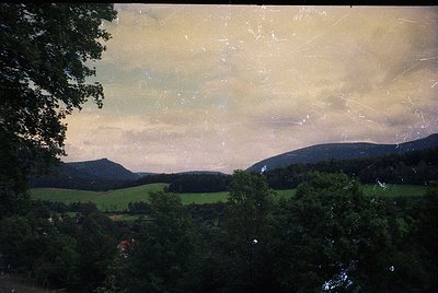 Vintage landscape photograph featuring rolling hills, dense forest, and a misty horizon. The faded, sepia-toned print suggest...