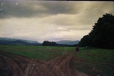 Vintage landscape shot of a rural dirt road winding through lush green fields under overcast skies. Two figures in dark cloth...