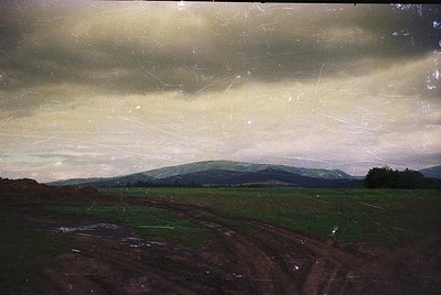 Vintage landscape photo showing rolling hills, farmland, and a cloudy sky. Evidence of aging film grain and scratches. Likely...