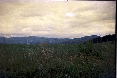 Vintage landscape shot featuring rolling hills under a dramatic, cloud-laden sky. Golden light filters through haze, creating...