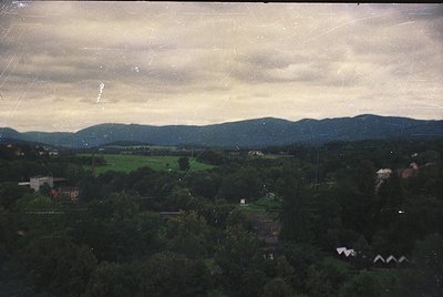 Vintage aerial view of rural landscape with dense forest, rolling hills, and scattered buildings. Overcast sky with visible s...