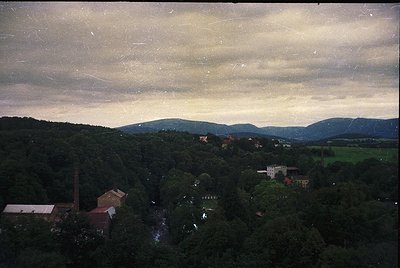 Vintage aerial view of a forested valley with industrial buildings and scattered structures. Dense tree cover dominates the l...