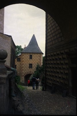Medieval-style courtyard framed by arched stone walls, featuring a small tower with conical roof and brickwork. Two figures i...
