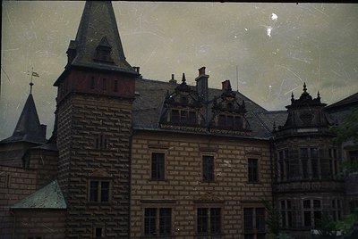 Vintage brick building with Gothic Revival architecture—steep gables, ornate dormer windows, and a corner turret. Yellowed, s...