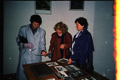 Three individuals examine framed photographs on a display table in a dimly lit indoor setting, likely a museum or gallery. Th...