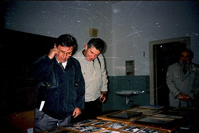 Three men examine framed black-and-white photographs in a dimly lit room, likely a museum or archive. The man on the left wea...
