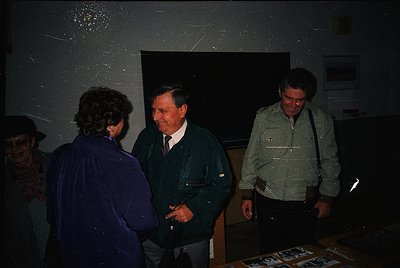 Indoor gathering featuring three men in 1970s-era attire—dark blazers, collared shirts, and shoulder bags—engaged in conversa...