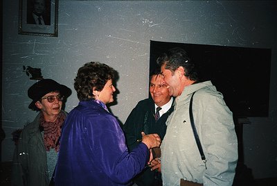 Indoor gathering featuring four adults in mid-1990s attire. A woman in a purple jacket shakes hands with a man in a light jac...
