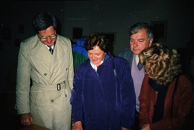 Four individuals in 1970s-era attire—men in blazers, women in raincoats—pose indoors under dim lighting. The man on the left ...