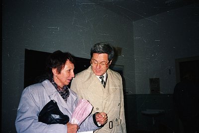 Two individuals in formal attire converse indoors, likely in a public or institutional setting. The woman wears a light gray ...