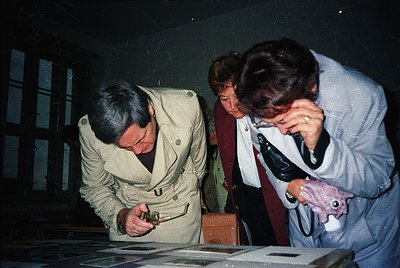Three individuals examine vintage photographs in a dimly lit room, likely a museum or archive. The man in the center wears a ...