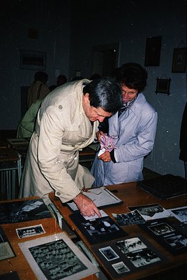 Two men examine vintage black-and-white photographs in a museum display case, likely from the 20th century. The man in the fo...