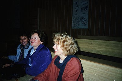 Three women in 1970s-style attire—blazers, scarves, and curly hair—seated indoors under dim lighting. Wooden paneling and a p...