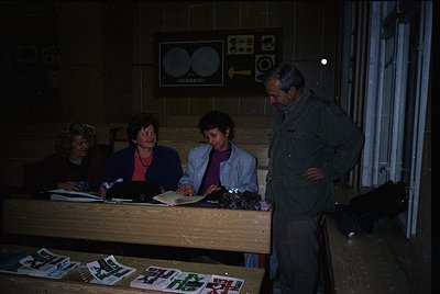 Four individuals in a dimly lit indoor setting, likely a 1970s-80s office or archive. Two women seated at a wooden desk revie...