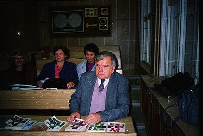 Indoor office setting from the 1970s-1980s, featuring a man in a suit and tie reviewing printed materials on a wooden desk. B...