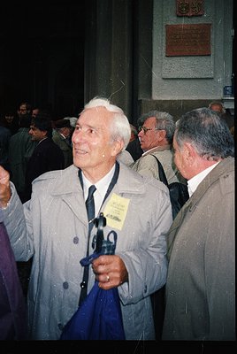 Mid-20th century formal event with elderly man in light coat, holding a blue umbrella and lanyard badge. Crowded indoor setti...