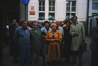 Group portrait in a European courtyard, likely 1970s–1980s. Eight individuals pose in vintage coats, scarves, and glasses, st...