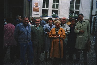 Group of 10 adults posing outdoors in 1970s European urban setting. Central woman in mustard-colored dress, surrounded by men...
