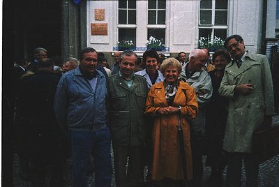 Group of seven adults posing in front of a European courtyard building, likely 1970s–1980s. Men wear jackets, ties, and overc...