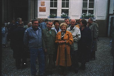 Group photo in a European courtyard, likely 1970s–1980s. Cobblestone ground and brick buildings with white-framed windows fra...