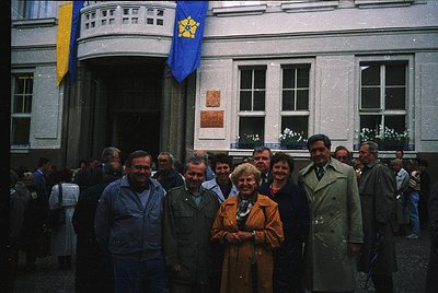 Neatly arranged group photo in front of a Soviet-era building, featuring a yellow sunburst emblem on a blue flag. Eight men a...