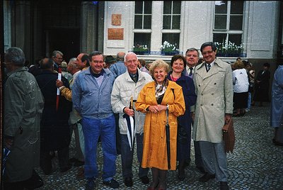 Group portrait in front of a neoclassical building with cobblestone courtyard, likely 1970s–1980s. Six adults pose in vintage...
