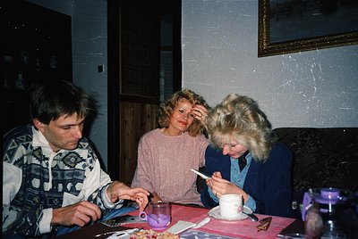 Three individuals seated indoors, likely mid-1990s, in a cozy, textured-wall setting. Left: man in patterned sweater, holding...