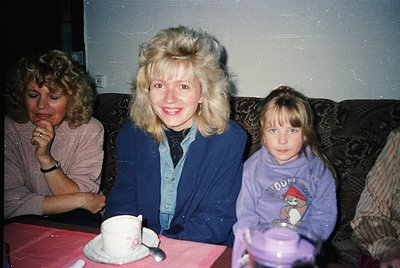 Three women pose indoors, likely 1980s–1990s. Center: woman in a blue cardigan with voluminous hair, smiling. Left: seated wo...