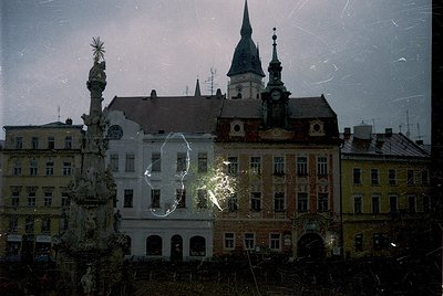 Vintage urban architecture through rain-spattered glass, featuring a Baroque-style fountain with ornate spire and clock tower...