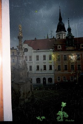 Vintage photograph of a European cityscape viewed through a rain-streaked window. Central column with ornate golden statue at...