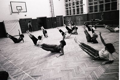 Group fitness routine in a gymnasium, likely 1960s–1980s. Participants perform floor exercises in unison, with one instructor...