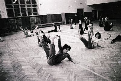 Group fitness class in a gymnasium, likely 1970s–1980s. Participants perform floor exercises in a spacious, wood-floored hall...