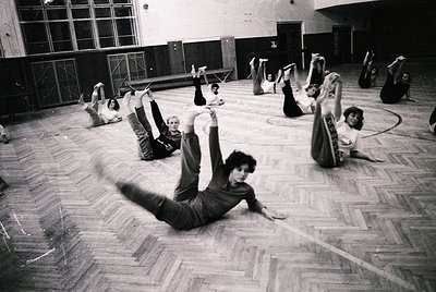 Group exercise class in a large indoor gymnasium, likely 1970s–1980s. Participants perform floor stretches in coordinated pos...