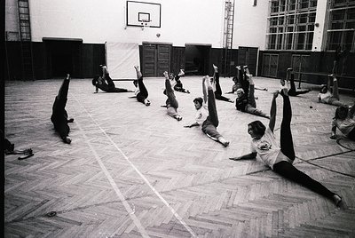 Group fitness class in a gymnasium, likely 1970s–1980s. Participants perform floor exercises in a large, wood-floored hall wi...