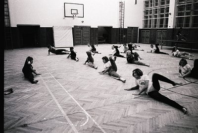 Indoor gymnasium scene with 15+ individuals performing floor exercises, likely a 1960s-1970s physical education class. Wooden...